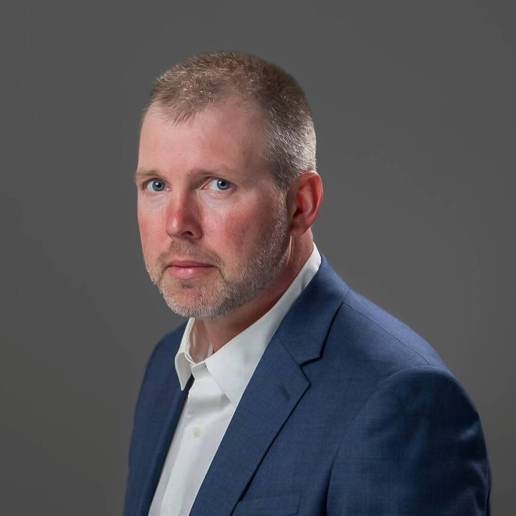 Professional headshot of Colin wearing a blue suit jacket and white shirt, looking at the camera with a neutral expression against a plain gray background